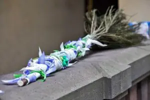 Ein Handfastingband, geknüpft aus verschiedenen Bändern in lila, weiß und grün mit handschriftlichen Wünschen, um einen Reisigbesen gewickelt liegt auf einer Steinmauer.