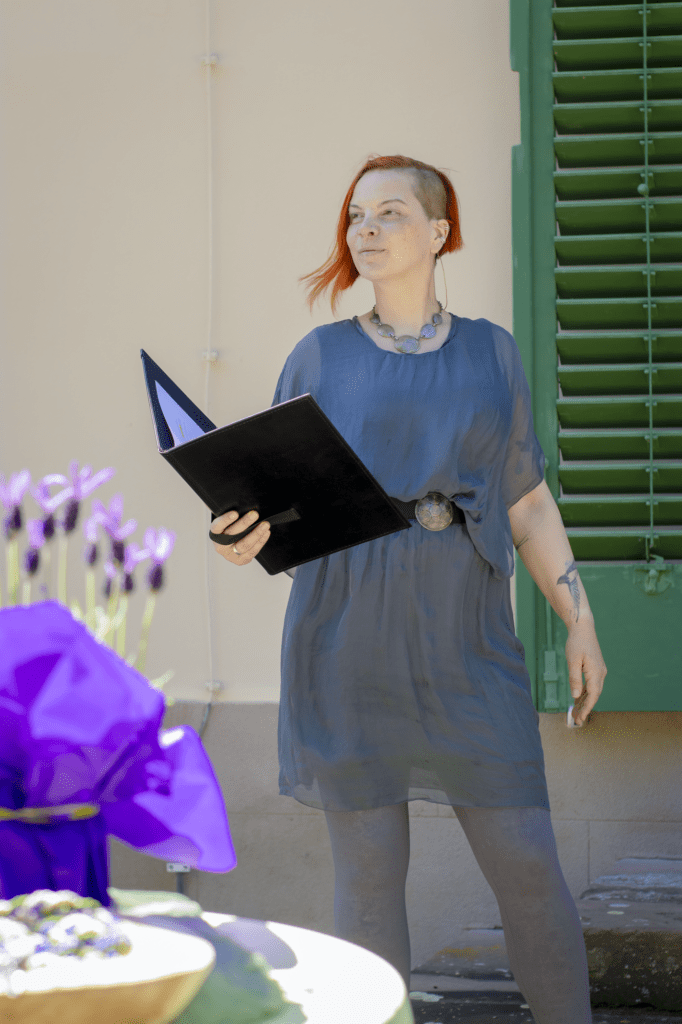 Friederike Amara Sonnenberg steht in einem grauen Seidenkleid mit aufgeschlagener Rednerinnenmappe und wartet mit einem Lächeln auf das Hochzeitspaar.